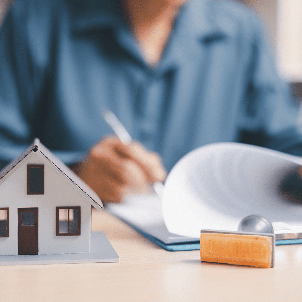 A person in a blue shirt fills out paperwork at a desk with a small model house and an orange "APPROVED" stamp in the foreground.
