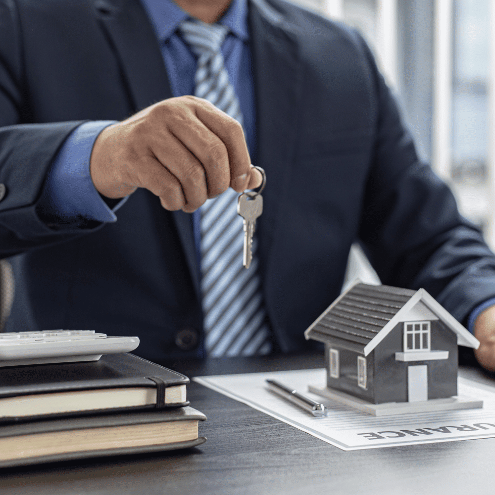 A person in a suit holds house keys over a table with a small model house, documents labeled "INSURANCE," books, a pen, and a calculator.