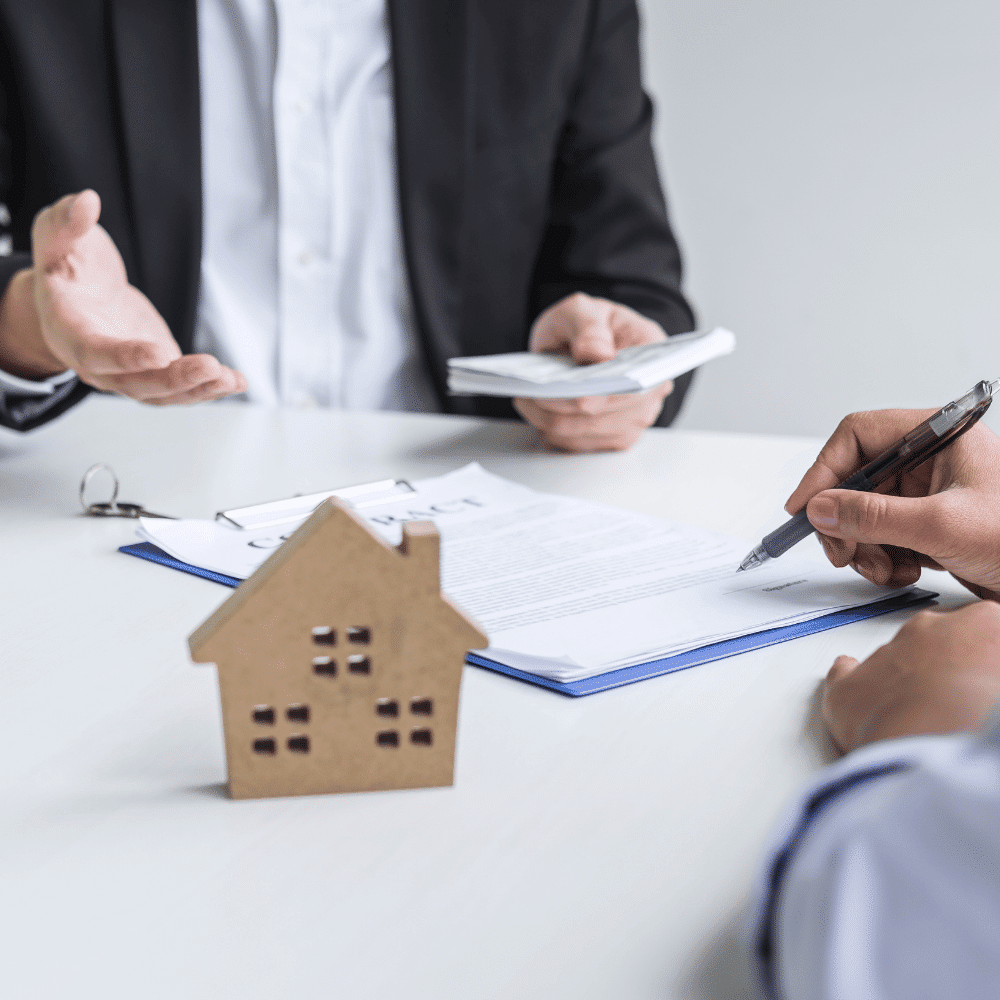 Two people sit at a table discussing documents related to a contract, with one person holding a pen and the other holding papers. A small wooden house model is on the table, suggesting a real estate transaction.