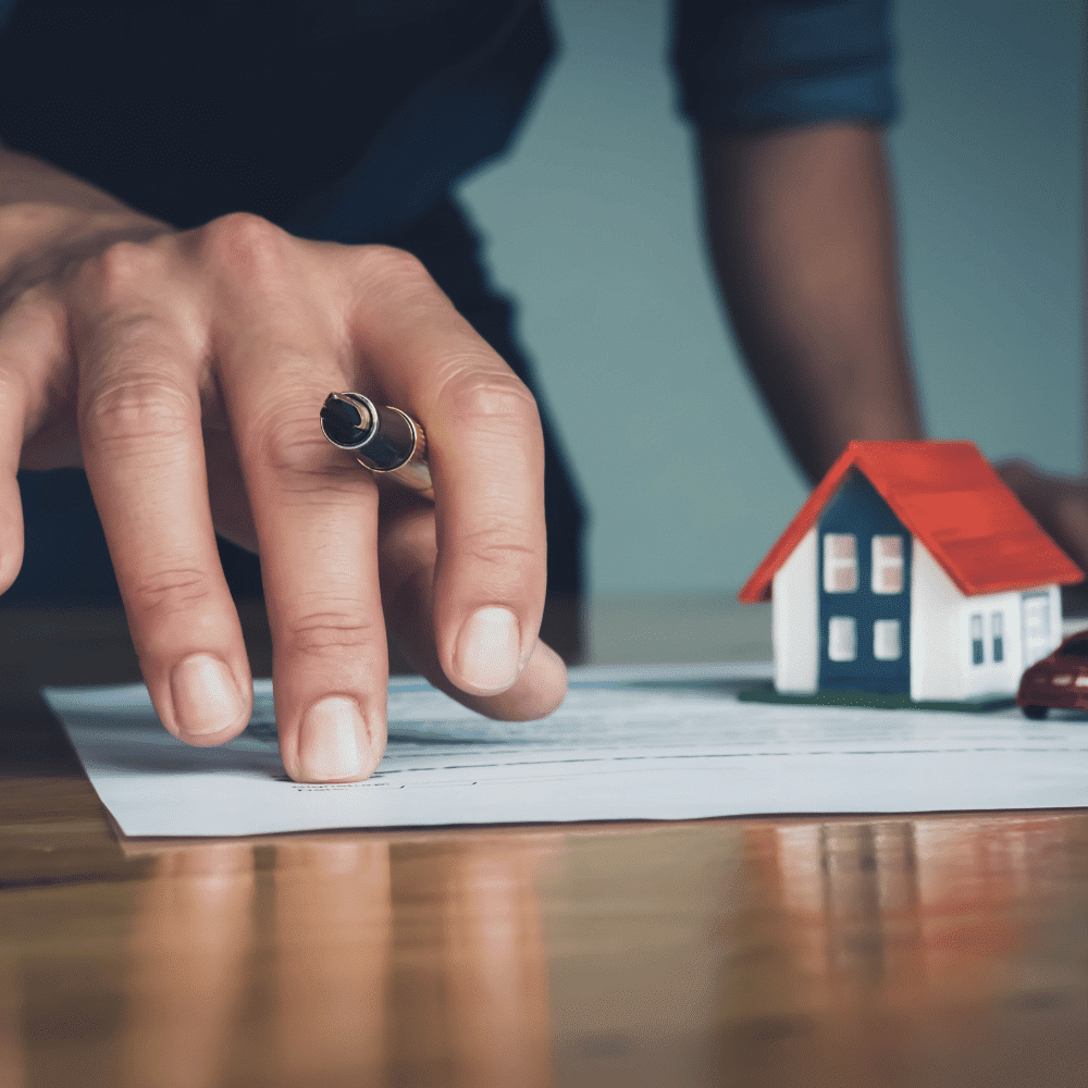 A person holding a pen is about to sign a document on a desk, with a small model house and a toy car nearby, symbolizing a real estate or mortgage agreement.