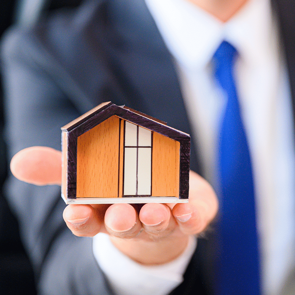 A person in a suit and blue tie holds a small wooden house model in their outstretched hand, symbolizing real estate, home ownership, or property investment.