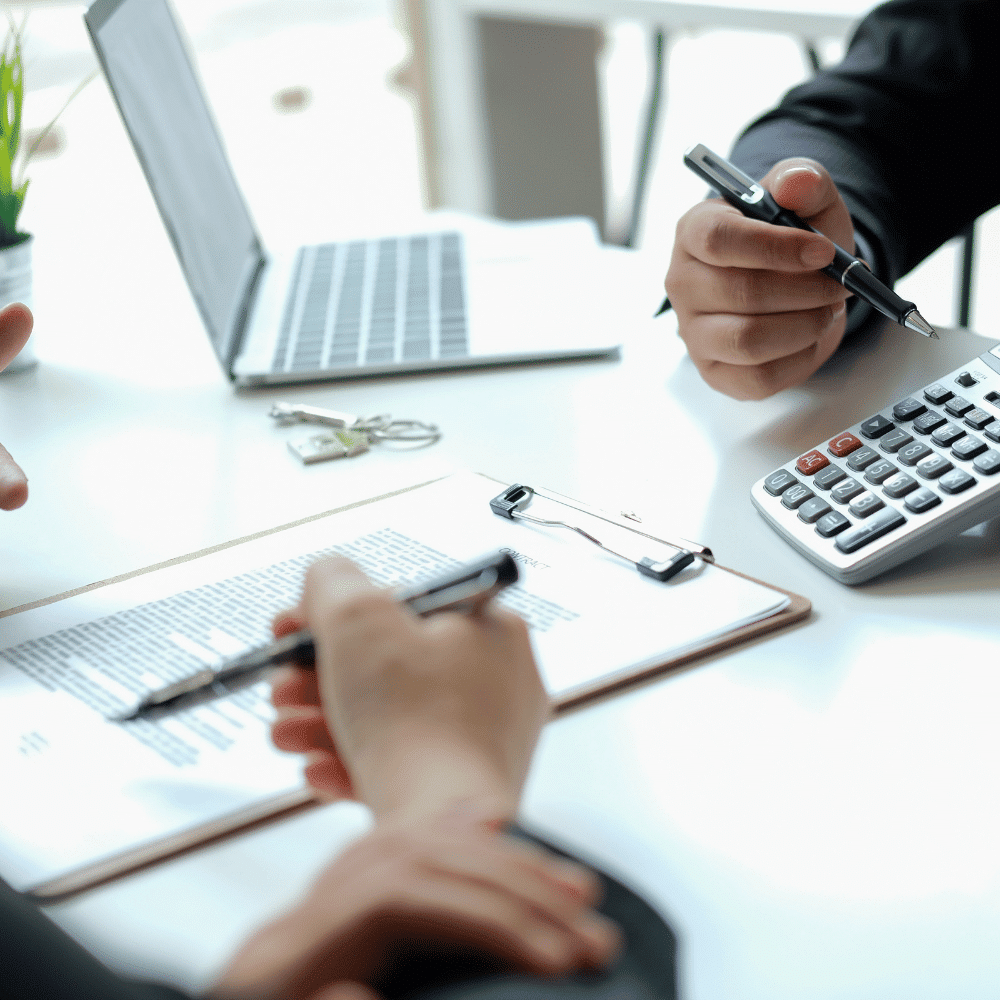 Two people sit at a desk with a clipboard, calculator, laptop, and keys, discussing documents and making calculations, suggesting a business or financial meeting.